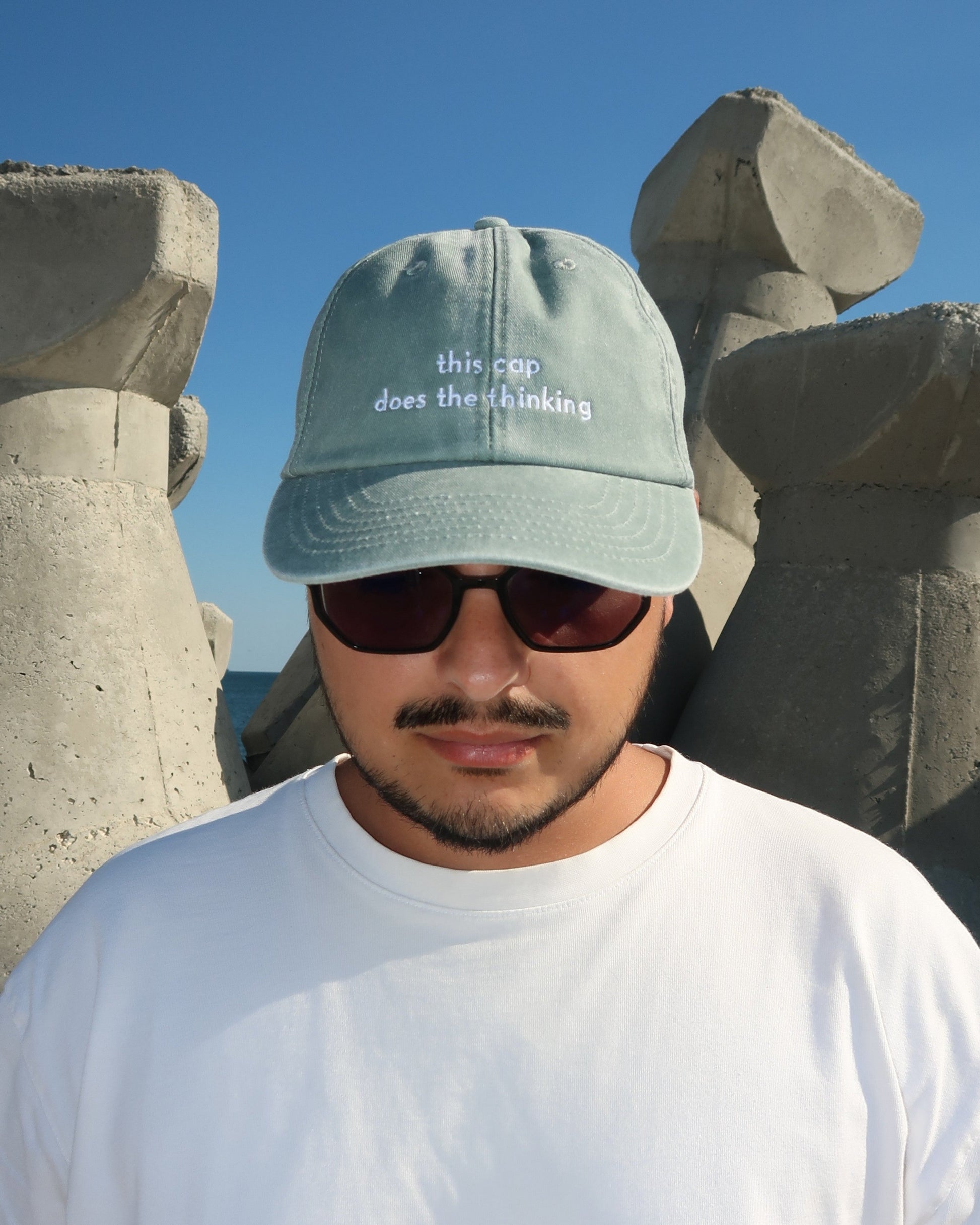 Person wearing a cool cap with text and sunglasses, standing against stone structures and blue sky.